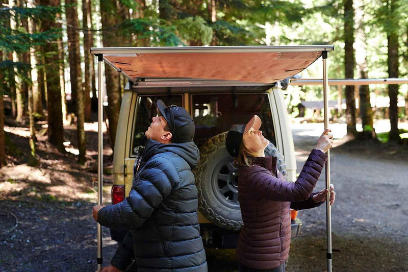Two campers setting the telescopic legs of a deployed ARB 814301 soft case awning beside a cream Toyota Land Cruiser in a forest