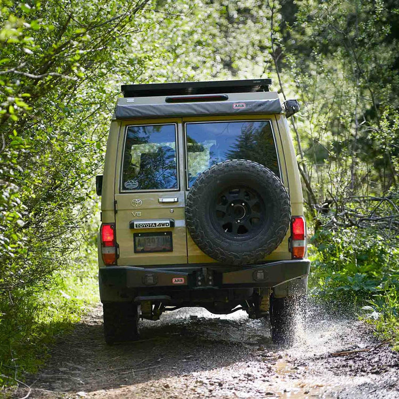 Beige Toyota 70-Series Land Cruiser on a forest trail carrying the packed ARB 814301 awning in its grey ARB Touring bag on the roof rack