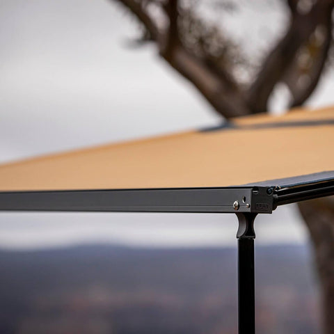 Detail of the ARB Hard Case Awning 814412A black alloy front rail, ARB-embossed cast leg joint and sand canopy while deployed outdoors