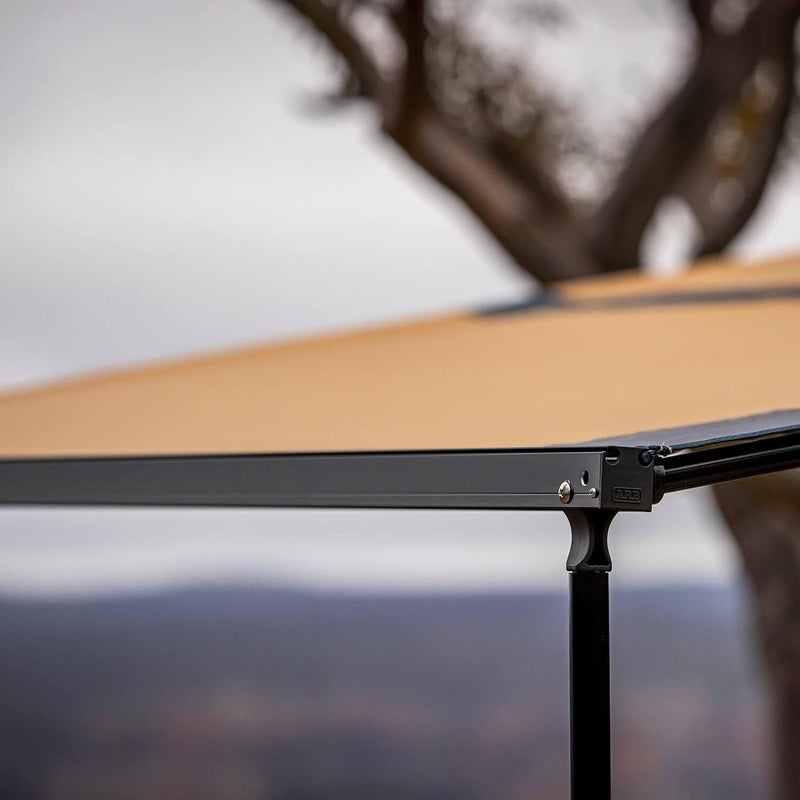 Detail of the ARB Hard Case Awning 814412A black alloy front rail, ARB-embossed cast leg joint and sand canopy while deployed outdoors