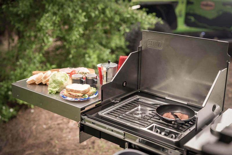 ARB Slide Out Camp Kitchen RDKNA1045 in use with a steak cooking on the burner and food prepared across the extended stainless bench