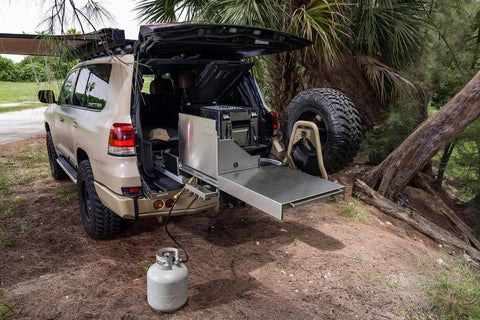 ARB Slide Out Camp Kitchen RDKNA1045 extended from the rear of a tan Land Cruiser, stainless bench deployed with a propane bottle connected