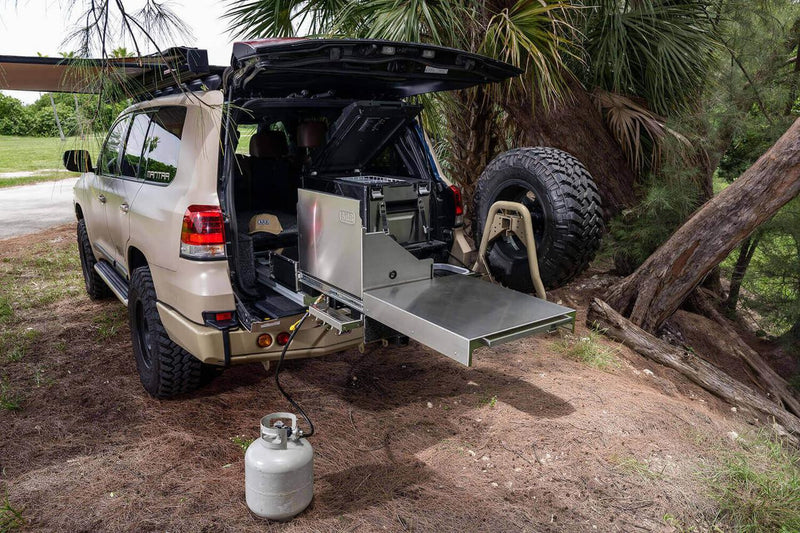 ARB Slide Out Camp Kitchen RDKNA1045 extended from the rear of a tan Land Cruiser, stainless bench deployed with a propane bottle connected