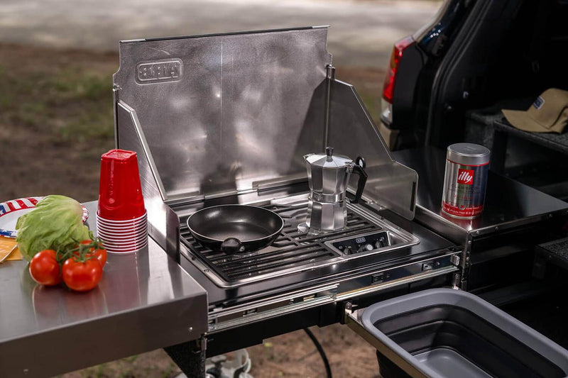 ARB Slide Out Camp Kitchen RDKNA1045 in use, stainless two-burner stove with ARB windshield raised and food laid out on the bench
