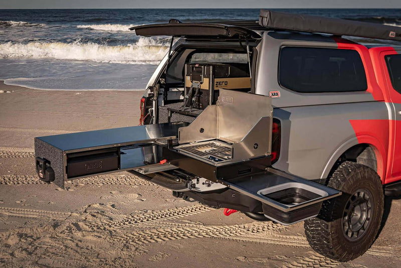 ARB Slide Out Camp Kitchen RDKNA1355 fully deployed from a truck tray on sand, showing the extended bench, stainless stove with wind shields and collapsible sink