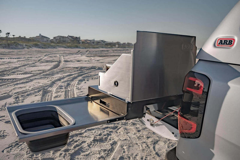 Rear quarter view of the ARB Slide Out Camp Kitchen RDKNA1355 on a white ute, showing the brushed stainless wind shields with ARB logo and the drop-down sink table