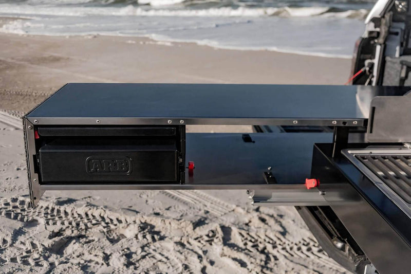 The black slide-out prep bench of the ARB Camp Kitchen RDKNA1355 extended from the tray, showing the ARB-embossed utensil drawer and red slide locking levers