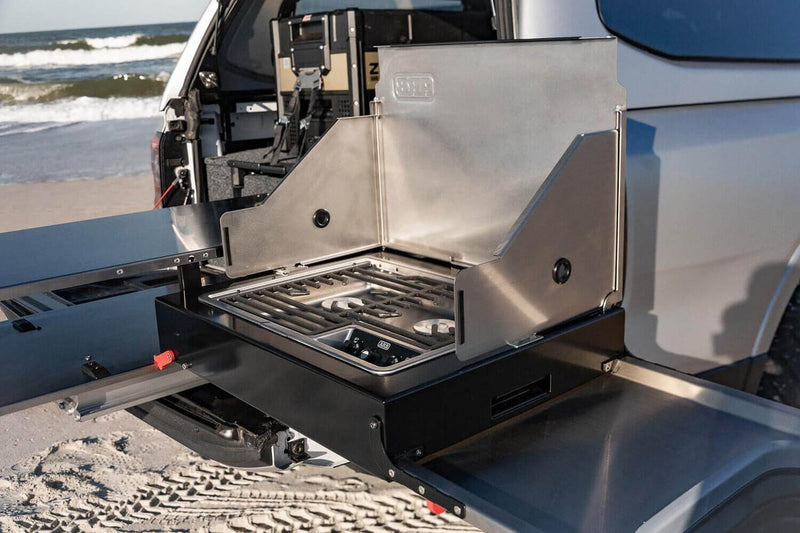 Side view of the ARB Slide Out Camp Kitchen RDKNA1355 stove module with wind shields raised, burner grate and controls, ARB ZERO fridge visible in the canopy