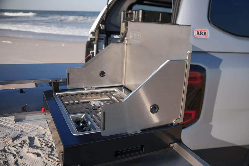 The ARB Slide Out Camp Kitchen RDKNA1355 stove seen along the deployed slide, with three-sided stainless wind shields raised around the burner grate