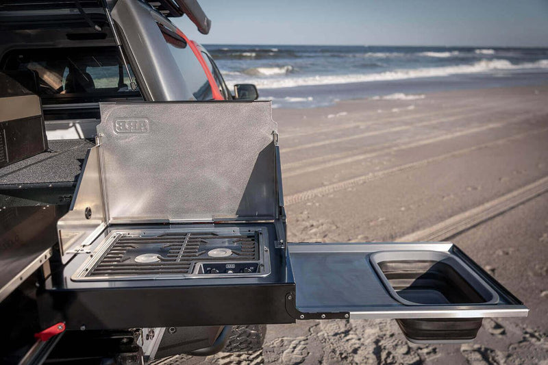 Close view of the ARB Slide Out Camp Kitchen RDKNA1355 two-burner stainless stove with wind shields raised and the collapsible silicone sink on the bench