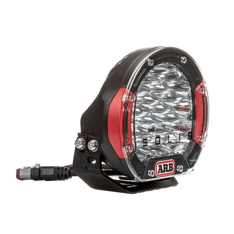 ARB Intensity Solis 21 flood driving light at an angle, squat black die-cast body with red side accents, clear lens over faceted flood optics and ARB INTENSITY SOLIS branding, with a sealed pigtail