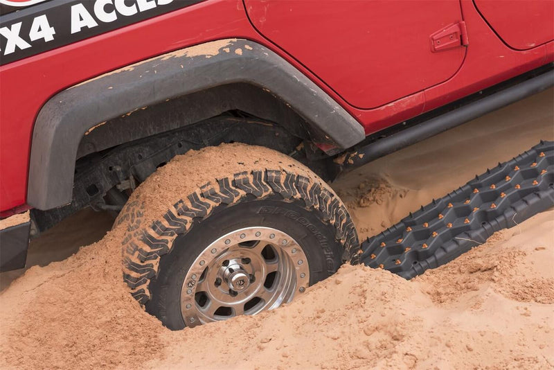 A pair of black TRED Pro recovery boards wedged under the rear tire of a red Jeep Wrangler bogged in desert sand