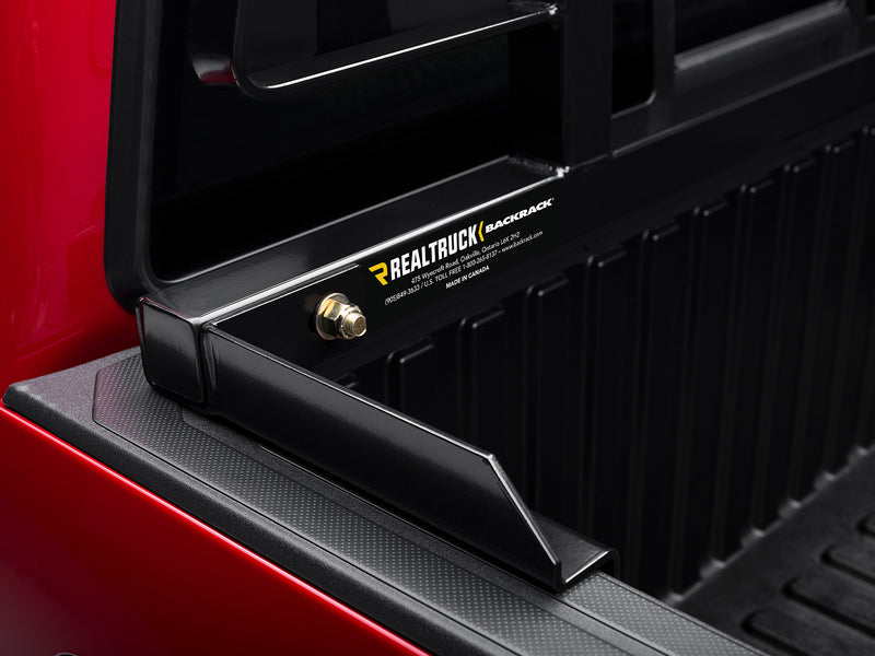 BackRack Original: close-up of a black headache rack base rail and bracket resting on a red pickup bed rail, showing the RealTruck BackRack label