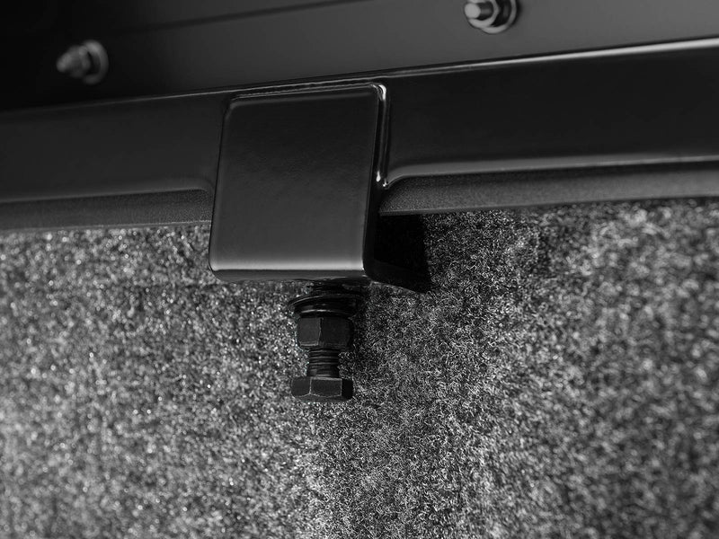 BackRack Trace Rack: close-up of a black clamp bracket and bolt fastened under a bed rail above a carpeted bed floor
