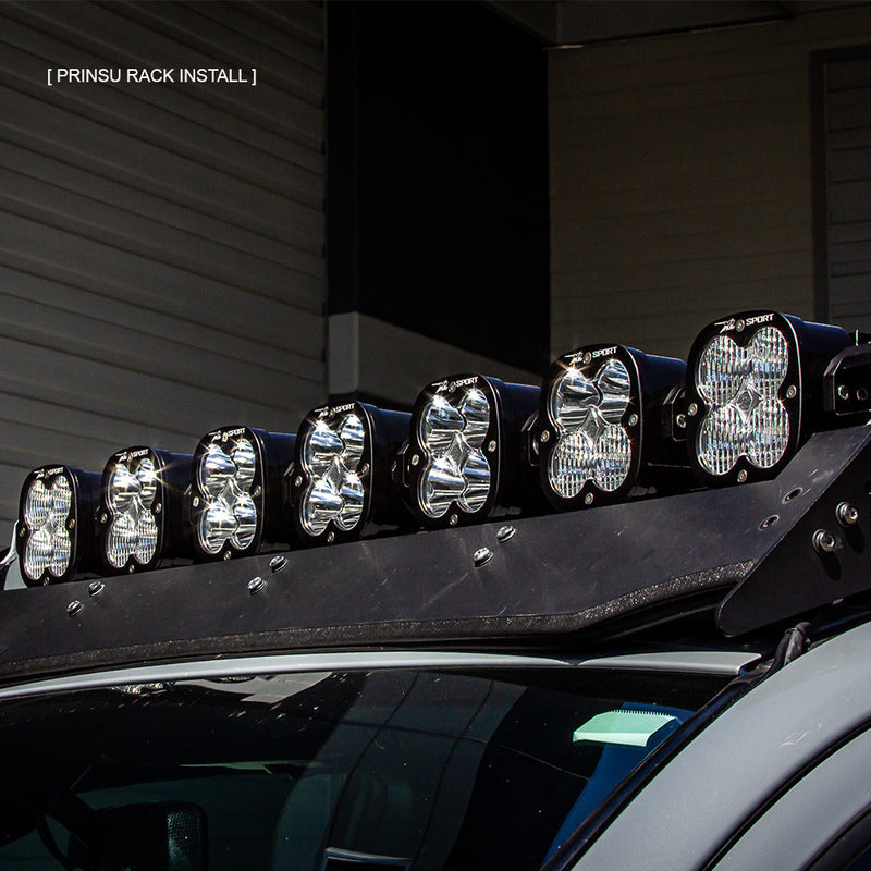 Baja Designs 7XL Linkable Roof Light Bar Kit For Prinsu Rack on a vehicle: seven linked XL pods mounted along the front edge of a black roof rack above a windshield