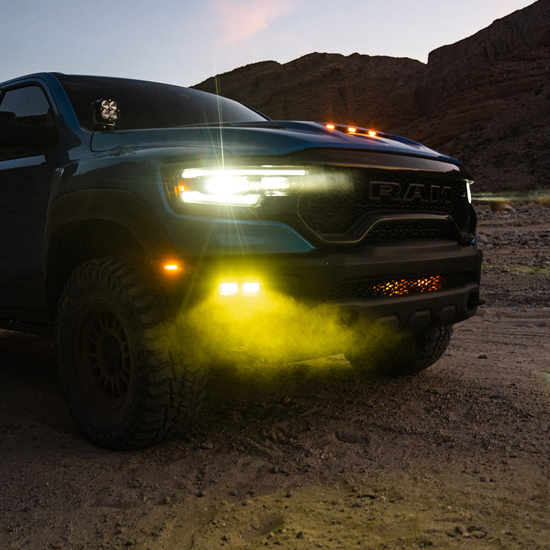 Baja Designs Dual S2 SAE Fog Pocket Light Kit installed on a RAM 1500 TRX: blue RAM 1500 TRX at dusk on a desert trail with amber fog lights glowing and a pod on the A-pillar