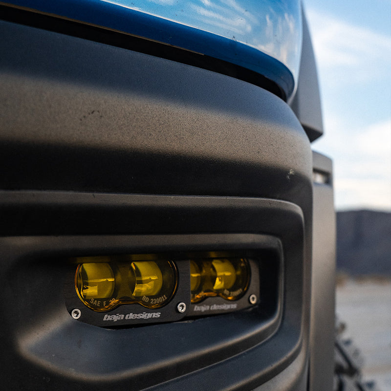 Baja Designs Dual S2 SAE Fog Pocket Light Kit on a vehicle: two S2 SAE pods with amber lenses mounted in the fog pocket of a dark blue truck bumper