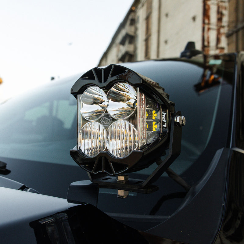 Baja Designs LP4 Pro LED Auxiliary Light Pod on a vehicle: A clear-lens LP4 Pro LED pod mounted on a hood-edge bracket of a dark vehicle in front of an old brick building