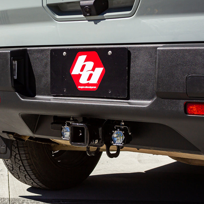 Baja Designs S1 Dual Reverse Light Kit on a vehicle: two S1 pods mounted on either side of a receiver hitch below a rear bumper with a Baja Designs logo plate