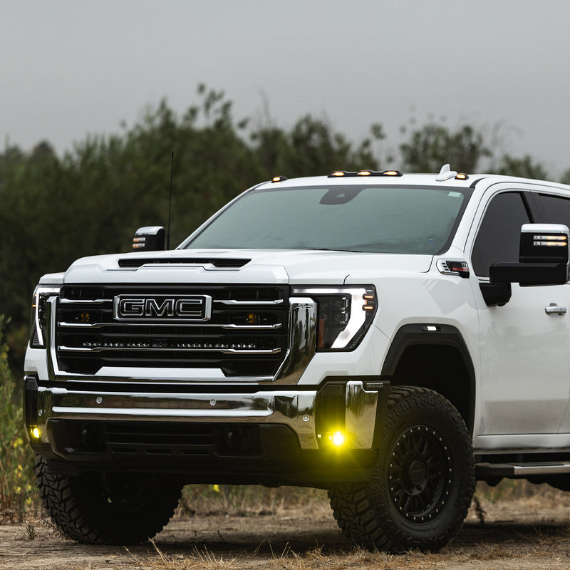 Baja Designs S1 Fog Pocket Light Kit installed on a GMC Sierra 2500HD/3500HD: A white GMC Sierra HD pickup parked on dirt with amber fog pocket lights glowing in the front bumper