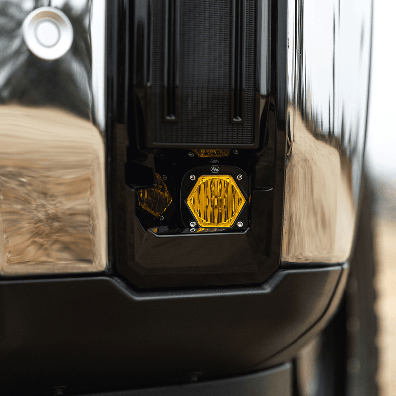Baja Designs S1 Fog Pocket Light Kit on a vehicle: close view of an amber S1 fog light glowing inside a black fog pocket next to a chrome bumper