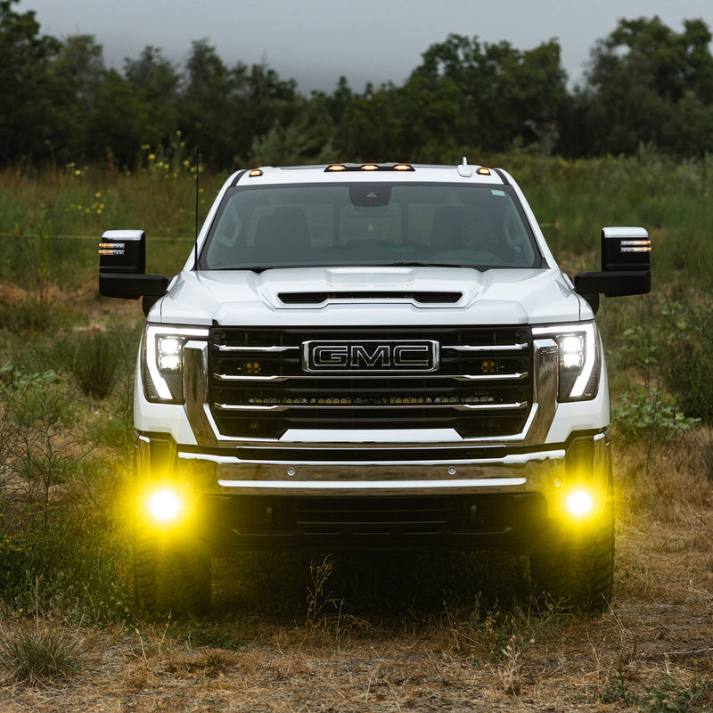Baja Designs S1 Fog Pocket Light Kit installed on a GMC Sierra 2500HD: white GMC Sierra HD parked in a field with lit amber fog lights in the front bumper