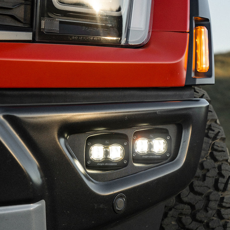 Baja Designs S2 SAE Dual Fog Pocket Kit on a Ford F-150 Raptor: close view of a red Ford Raptor front bumper with two lit S2 pods in the fog pocket