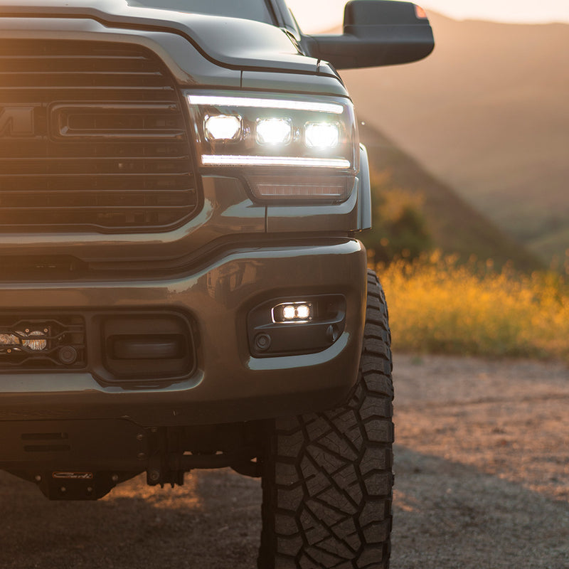 Baja Designs S2 SAE Fog Pocket Light Kit installed on a Ram 2500/3500: A gray Ram heavy duty pickup at sunset with S2 fog lights glowing in the lower bumper pockets