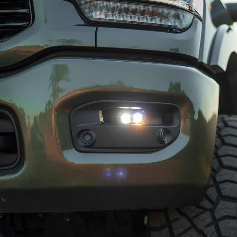 Baja Designs S2 SAE Fog Pocket Light Kit on a Ram 2500/3500: close view of a gray Ram heavy duty bumper with a lit S2 pod in the fog pocket under the headlight