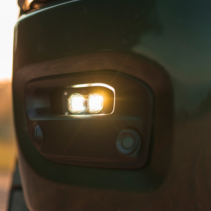 Baja Designs S2 SAE Fog Pocket Light Kit on a vehicle: close view of a lit S2 pod set into a dark truck fog pocket bezel at dusk