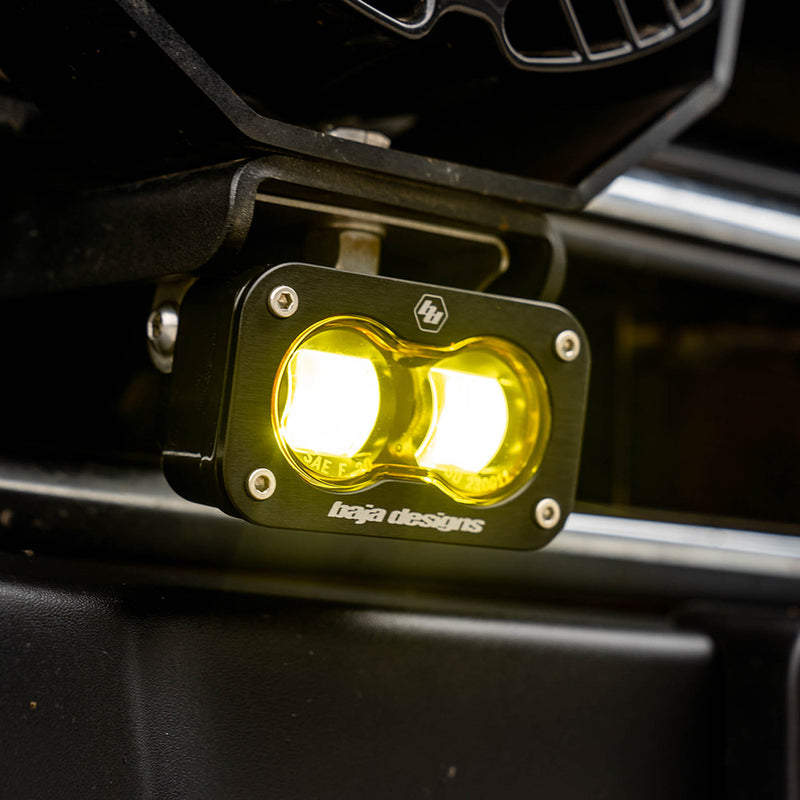 Baja Designs S2 SAE LED Auxiliary Light Pod Pair on a vehicle: A lit S2 SAE pod with an amber lens mounted on a bracket below a black vehicle grille
