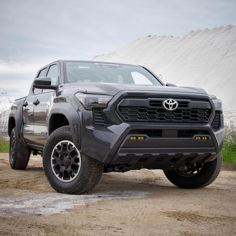 Baja Designs S2 SAE OEM Fog Light Replacement Kit installed on a Toyota Tacoma: A gray Toyota Tacoma pickup on a dirt lot with amber S2 pods fitted in its lower bumper fog pockets