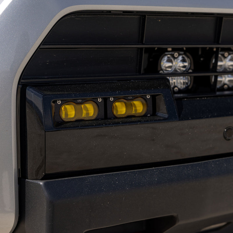 Baja Designs S2 SAE OEM Fog Light Replacement Kit on a vehicle: close view of two amber S2 fog lights set into a black lower bumper opening on a silver vehicle; amber shown while this frame is tied only to 44-8282, the clear SAE kit