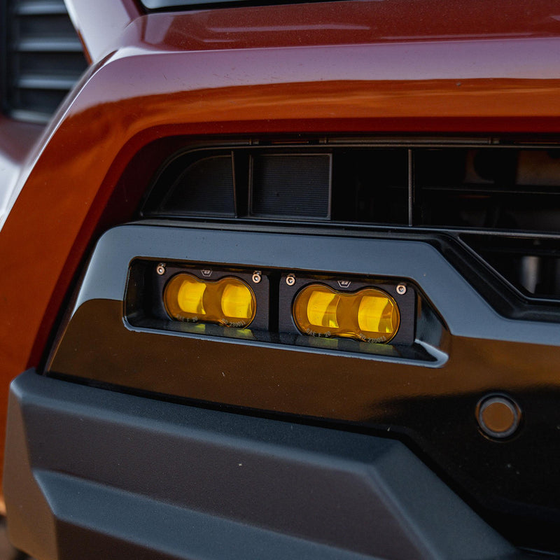 Baja Designs S2 SAE OEM Fog Light Replacement Kit on a vehicle: close view of two amber S2 fog lights set into the lower bumper opening of an orange vehicle