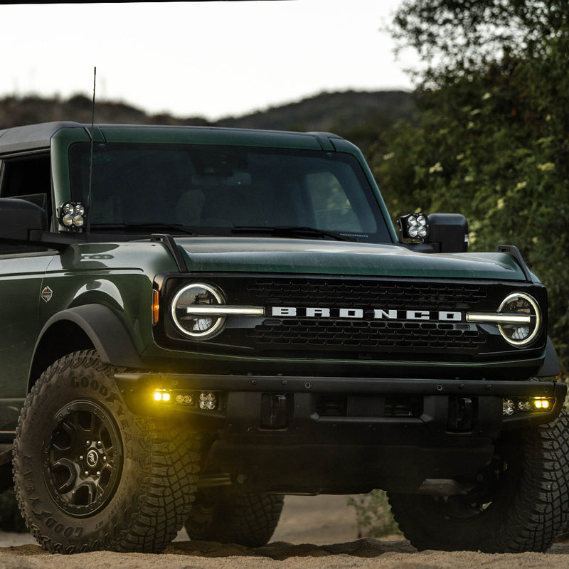 Baja Designs S2 SAE Pro Fog Pocket Light Kit installed on a Ford Bronco: A green Ford Bronco at dusk with amber fog pocket lights glowing in the steel front bumper