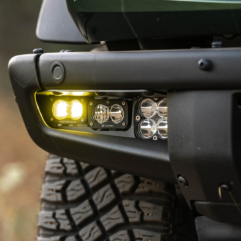 Baja Designs S2 SAE Pro Fog Pocket Light Kit on a vehicle: close view of a black steel bumper fog pocket with an amber S2 pod lit beside a clear S2 and a Squadron pod