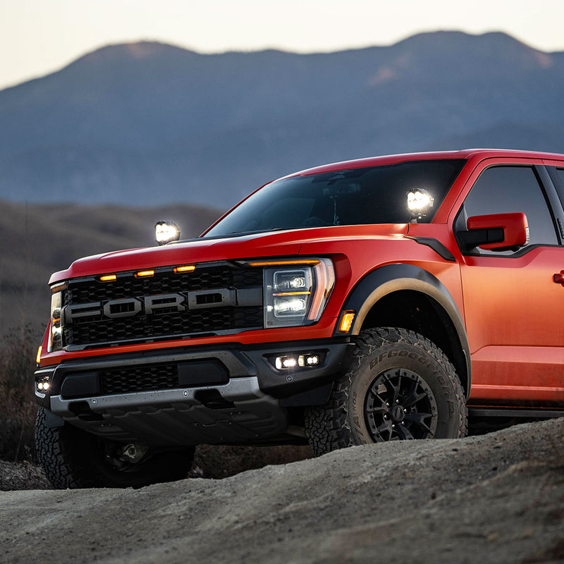 Baja Designs S2 SAE/S2 Pro Fog Pocket Kit installed on a Ford F-150 Raptor: A red Ford Raptor pickup parked on rocky ground at dusk with lit fog pocket lights in the lower bumper