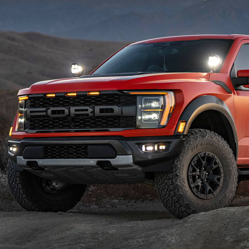 Baja Designs S2 SAE/S2 Sport Fog Pocket Kit installed on a Ford F-150 Raptor: A red Ford Raptor at dusk with lit fog pocket lights in the bumper and extra pods lit near the windshield