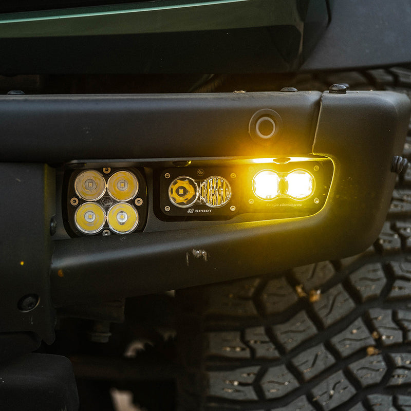 Baja Designs S2 SAE Sportsmen Fog Pocket Light Kit on a vehicle: close view of a black steel bumper fog pocket with a Squadron pod and two S2 pods glowing amber