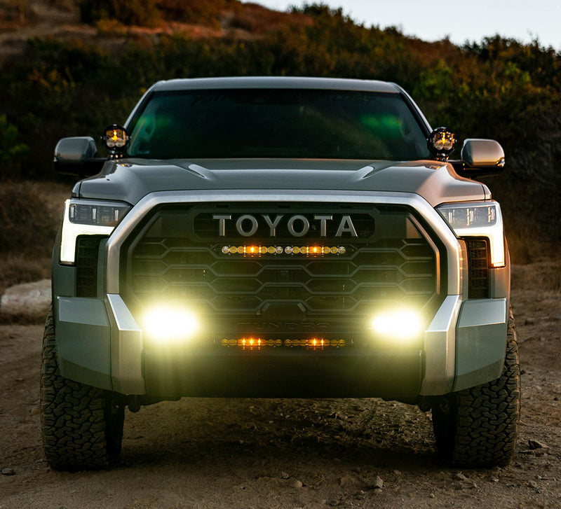 Baja Designs S2 Sport Dual Fog Pocket Light Kit installed on a Toyota Tundra: gray Toyota Tundra seen head-on at dusk with its fog pocket lights glowing and pods on the A-pillars