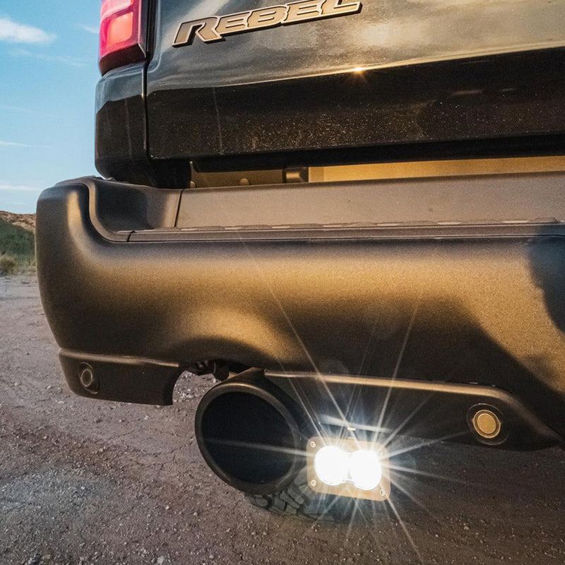 Baja Designs S2 Sport Dual Reverse Light Kit on a Ram 1500 Rebel: A lit reverse pod glowing under the rear bumper beside the exhaust tip of a Ram Rebel