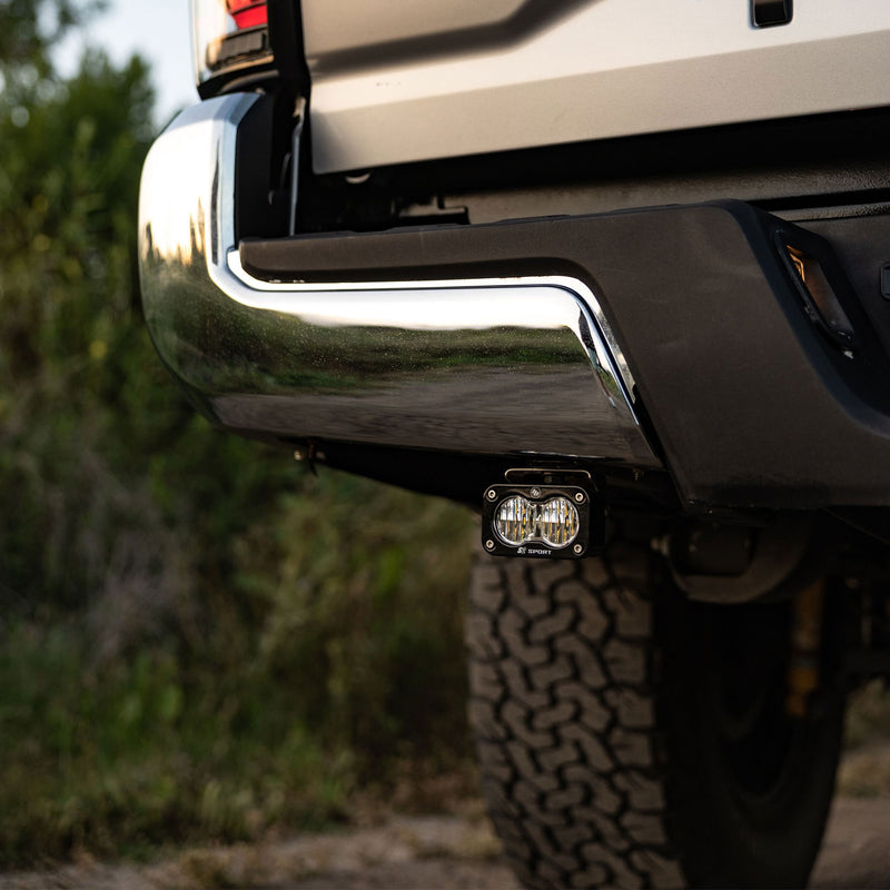 Baja Designs S2 Sport Dual Reverse Light Kit on a vehicle: an S2 pod mounted beneath a chrome rear bumper beside an off-road tire