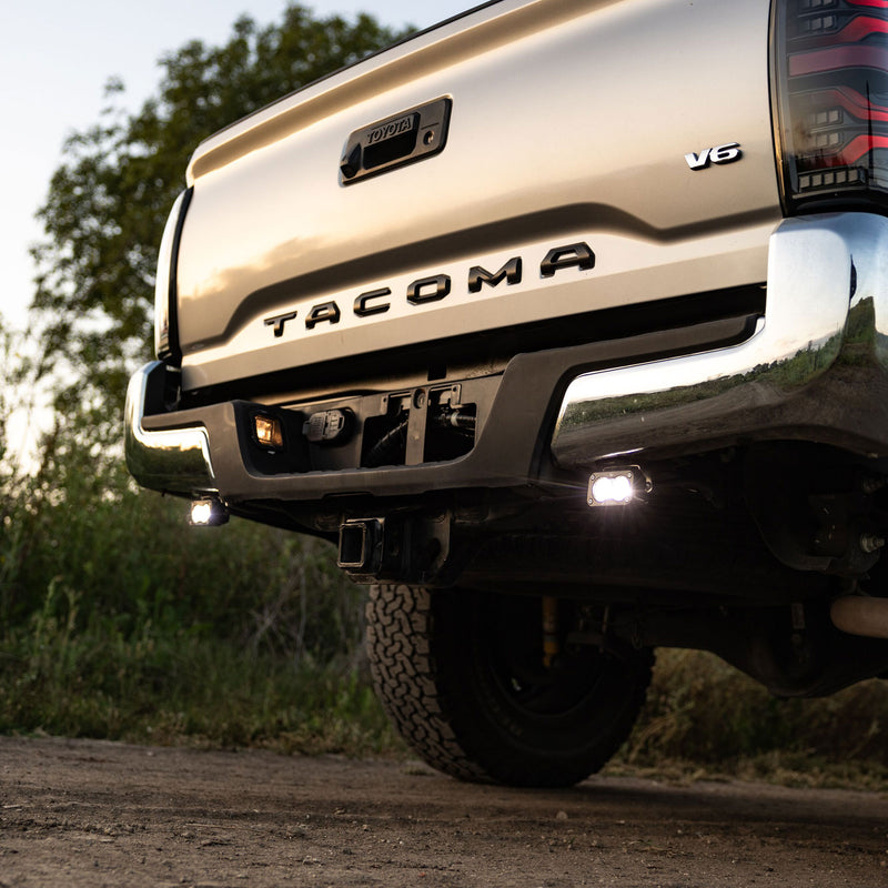 Baja Designs S2 Sport Dual Reverse Light Kit installed on a Toyota Tacoma: rear of a silver Toyota Tacoma on a dirt trail with two reverse pods glowing under the bumper