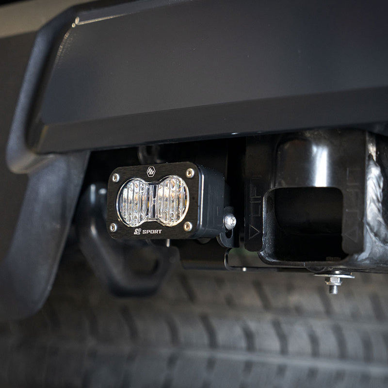 Baja Designs S2 Sport Reverse Kit on a vehicle: close view of a black S2 Sport LED pod mounted beside the receiver hitch under a black rear bumper