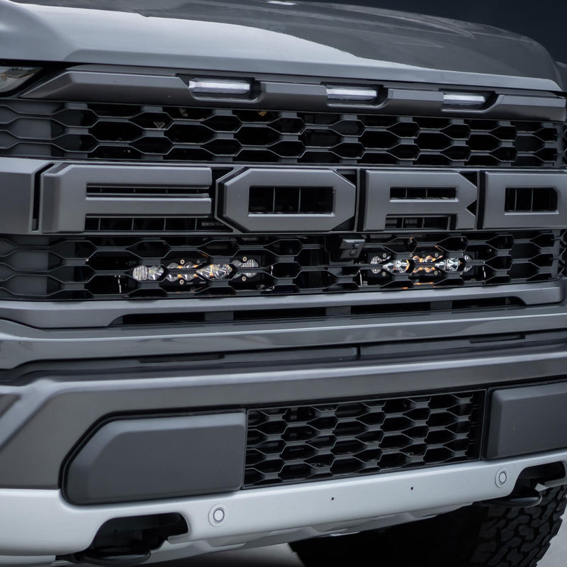 Baja Designs S8 10" Dual Behind Grille Light Bar Kit on a Ford F-150 Raptor: gray Ford Raptor grille with two short LED light bars mounted behind the lower grille below the FORD lettering