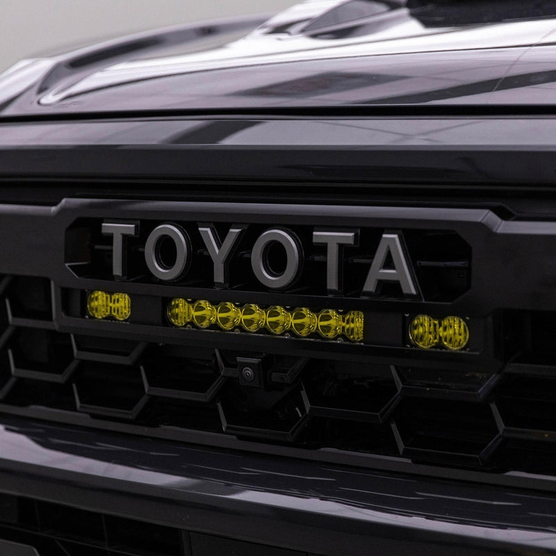 Baja Designs S8 10" Light Bar / S2 Pro TRD Pro Grille Conversion Light Kit on a Toyota: close view of a dark gray Toyota grille with an amber-lit S8 light bar and two S2 Pro pods beneath the TOYOTA lettering