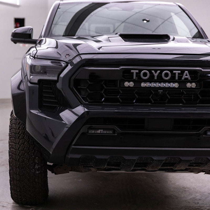 Baja Designs S8 10" Light Bar / S2 Pro TRD Pro Grille Conversion Light Kit installed on a Toyota Tacoma: front three-quarter view of a dark gray Toyota Tacoma in a studio with an S8 light bar and two S2 Pro pods in the grille