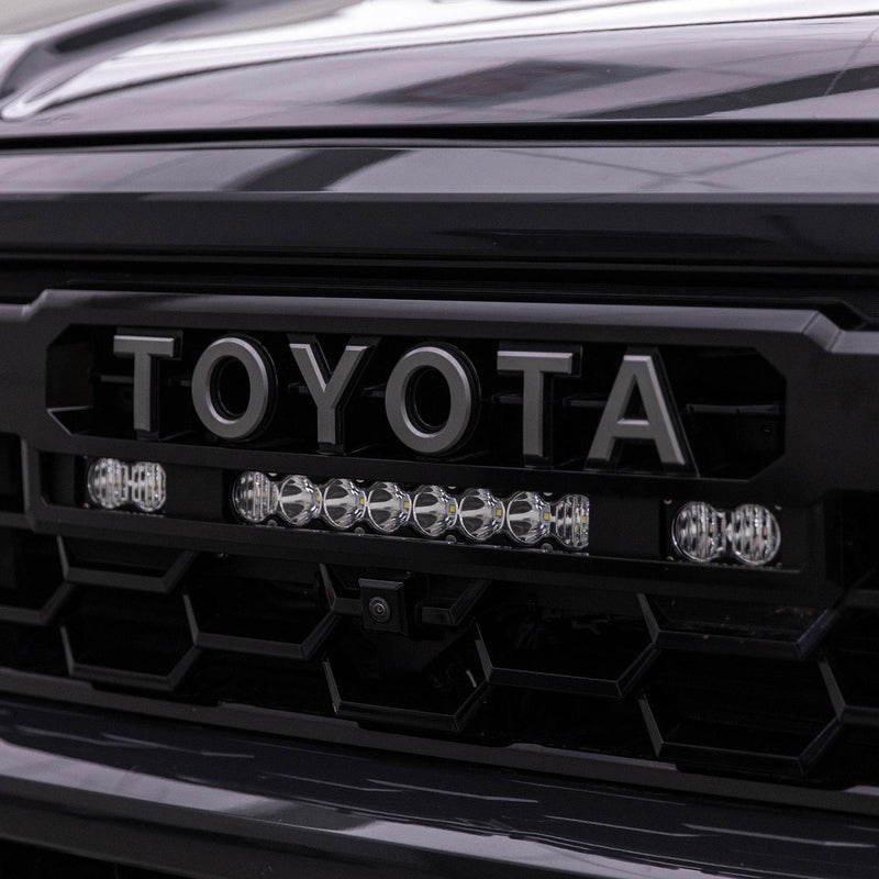 Baja Designs S8 10"/S2 Pro Grille Light Kit on a Toyota Tacoma: A clear lens 10" S8 bar between two S2 Pro pods mounted under the TOYOTA lettering of a dark Tacoma grille