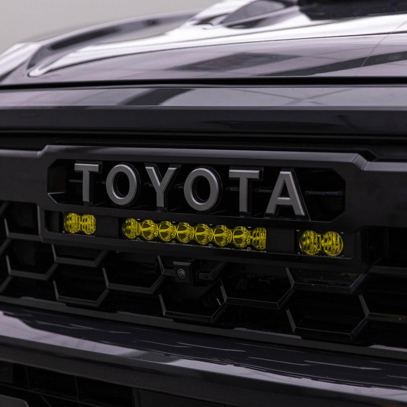 Baja Designs S8 10"/S2 Pro Grille Light Kit on a Toyota Tacoma: an amber 10" S8 bar between two amber S2 Pro pods sits under the TOYOTA lettering of a dark Tacoma grille