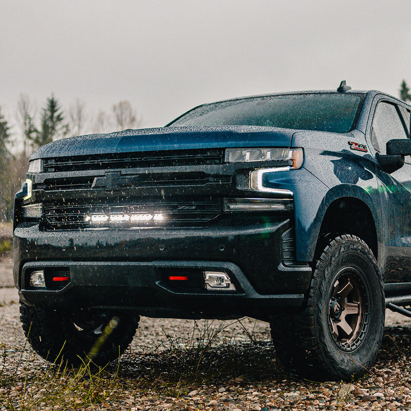 Baja Designs S8 20" Grille Light Bar Kit installed on a Chevrolet Silverado 1500: A dark blue Chevrolet Silverado parked on a gravel trail with a lit light bar glowing in the grille